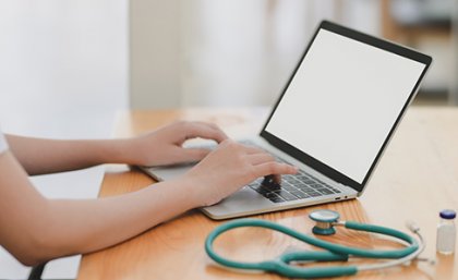 doctor typing on a laptop with a stethoscope curled about beside the keyboard, and a patient skyping or zooming for a telehealth consult 